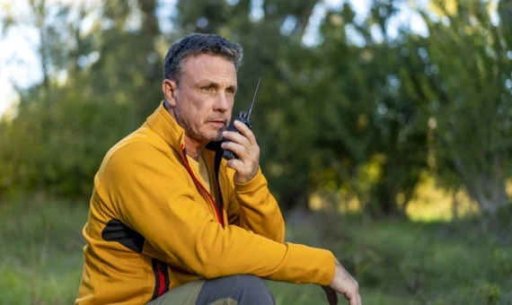 Man using a walkie talkie outdoors while seated in a forest clearing, wearing a yellow jacket during daytime
