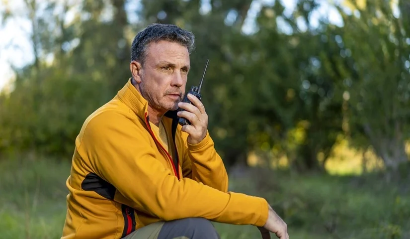 Man using a walkie talkie outdoors while seated in a forest clearing, wearing a yellow jacket during daytime