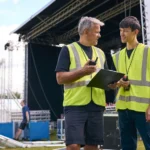 Two event staff in high-vis vests using walkie-talkies while coordinating at an outdoor stage setup.