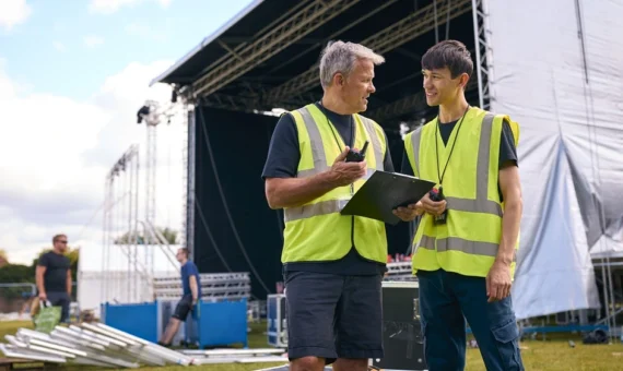 Two event staff in high-vis vests using walkie-talkies while coordinating at an outdoor stage setup.