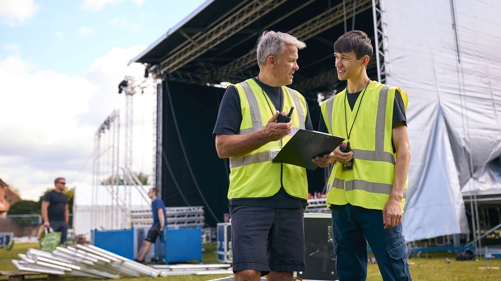 Two event staff in high-vis vests using walkie-talkies while coordinating at an outdoor stage setup.