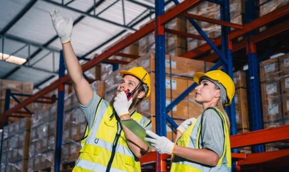 Warehouse workers in hi-vis vests and hard hats using a walkie-talkie and clipboard while checking stock on racking shelves.