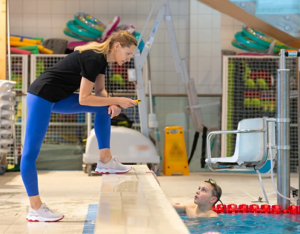 Leisure centre staff member using a walkie talkie beside an indoor pool while speaking to a child swimmer during a session.